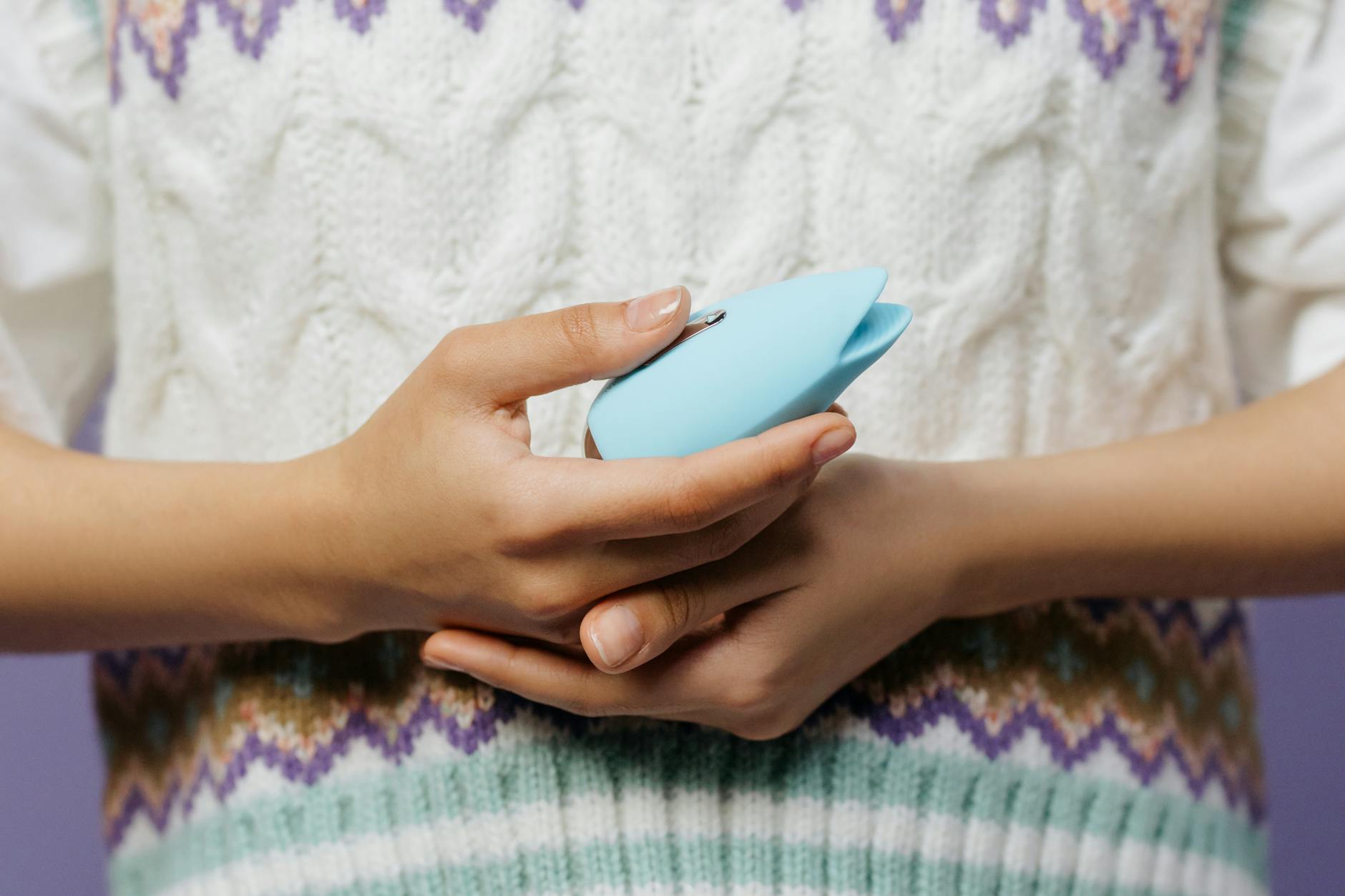 A hand holding a silicone clitoral vibrator against a knitted sweater, representing comfort and intimacy.